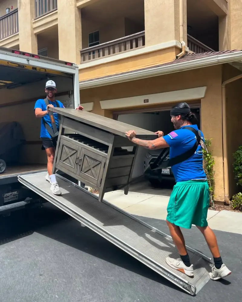 Two Flexdolly movers carrying a wooden cabinet down the truck ramp to a home