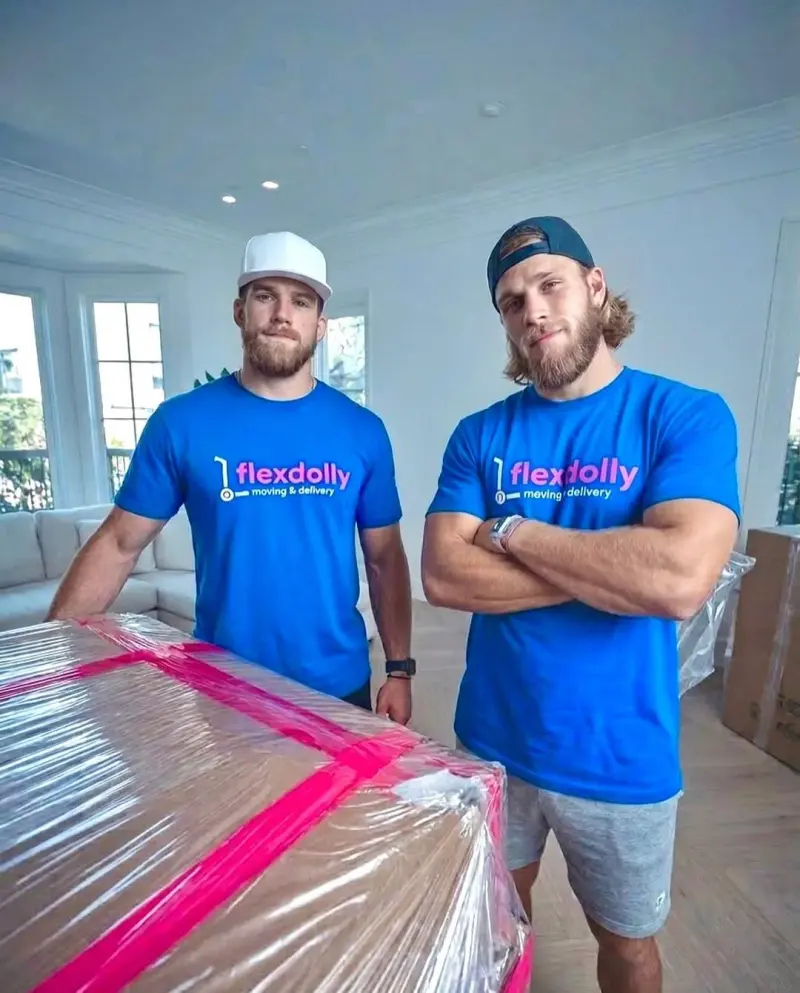Two Flexdolly crew members in a bright staged living room beside a large piece wrapped in protective film and pink tape